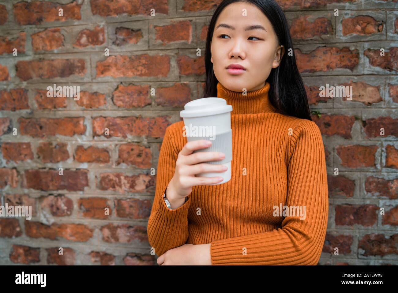 Portrait of young Asian woman holding a cup of coffee against brick ...