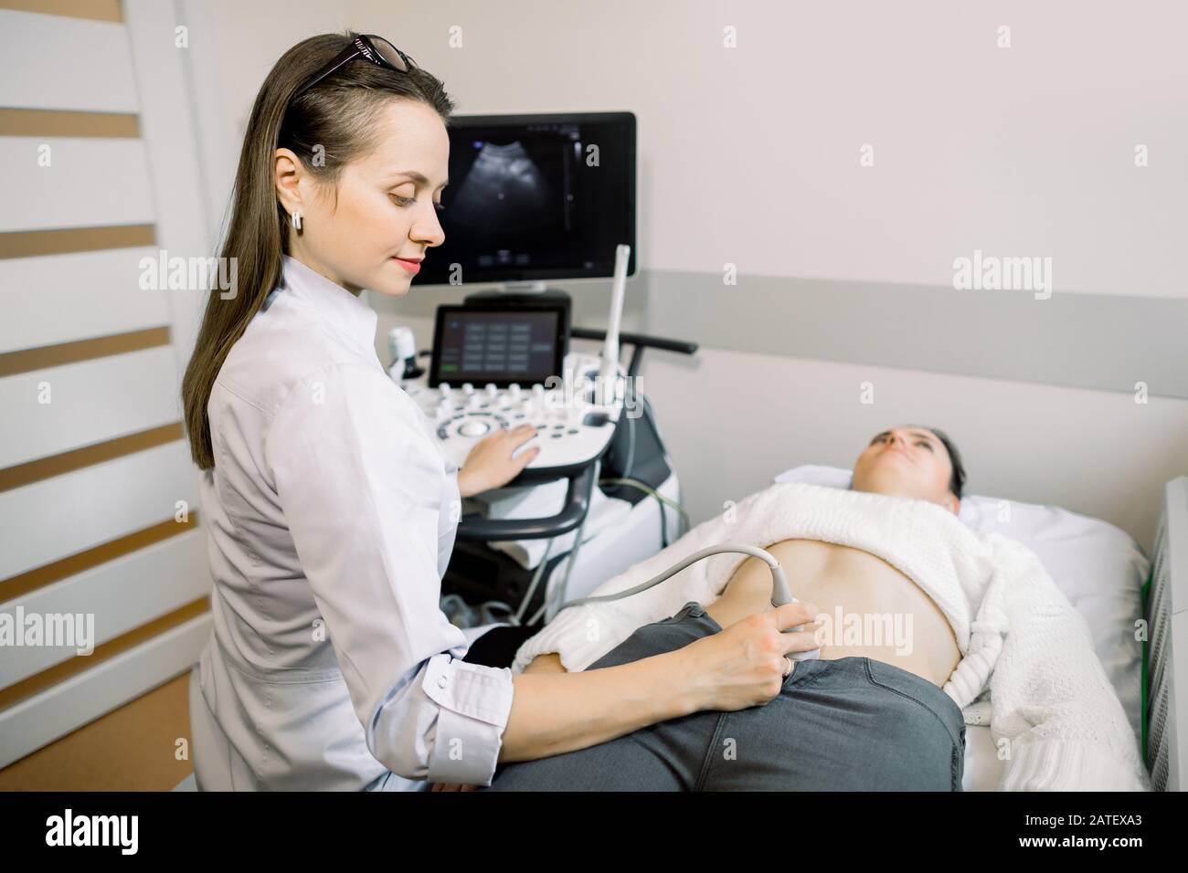 Pretty female doctor operating modern ultrasound scanner while examining belly and bladder of her female patient. Stock Photo