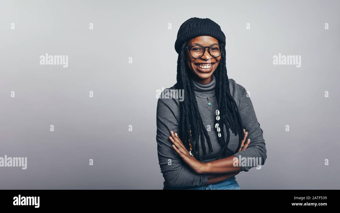 Portrait of cheerful african female having long dreadlocks looking at camera. Smiling african woman with her arms crossed isolated on grey background. Stock Photo
