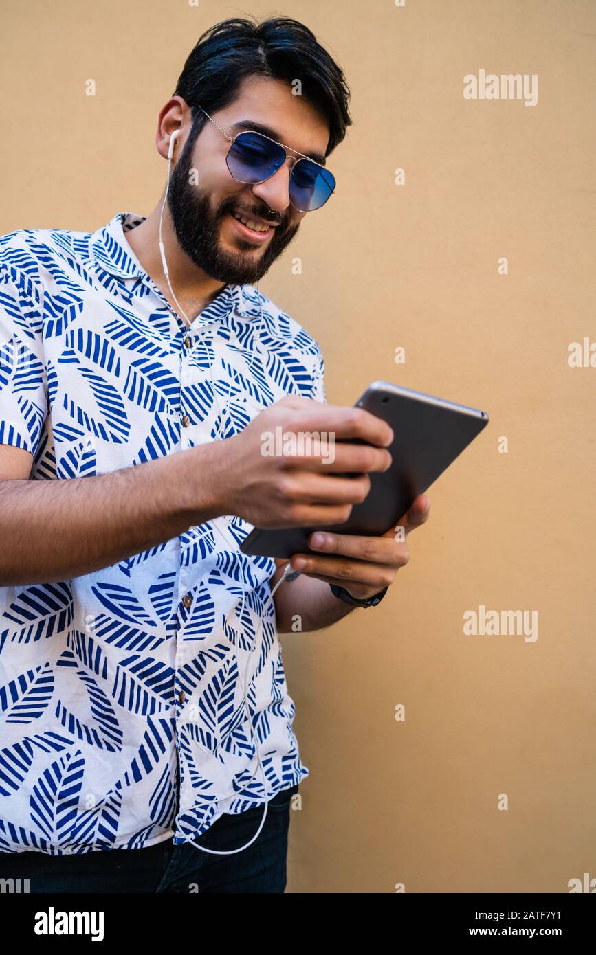 Portrait of young latin man using his digital tablet with earphones against yellow wall. Technology and urban concept. Stock Photo