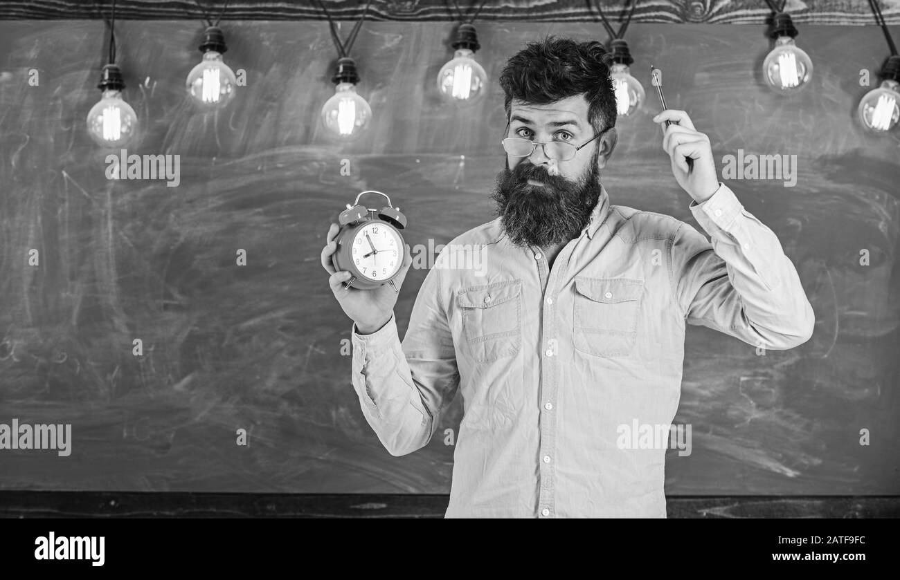 Man with beard and mustache on forgetful face itching head. Bearded hipster holds clock, chalkboard on background, copy space. Discipline concept. Teacher in eyeglasses holds alarm clock and pen. Stock Photo