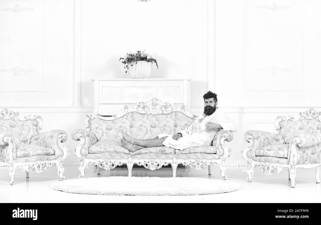 Man sleepy in bathrobe drinks coffee in luxury hotel in morning, white background. Luxury life concept. Man with beard and mustache enjoys morning while sitting on old fashioned luxury sofa. Stock Photo