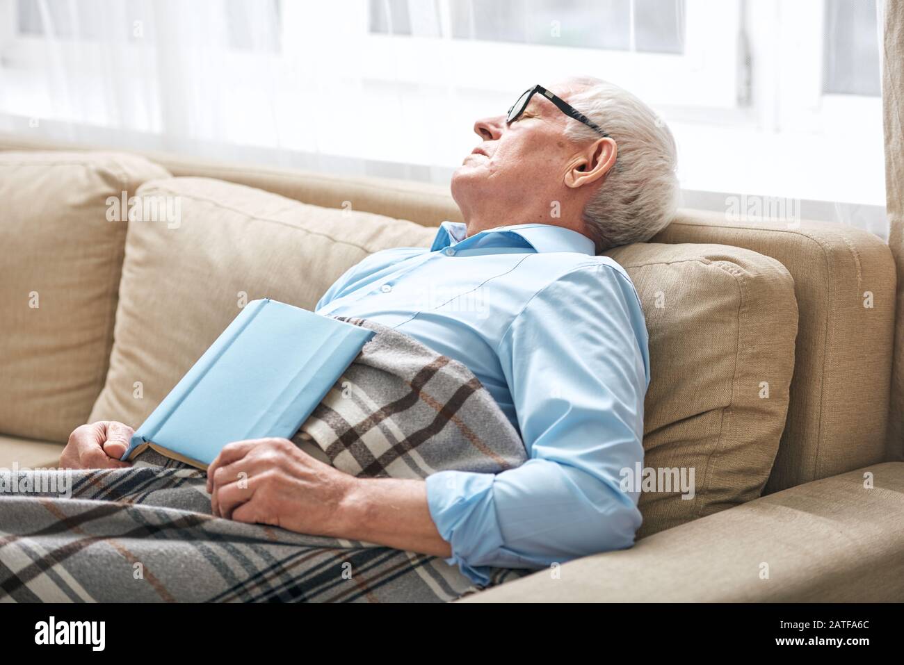 Tired senior man covered with plaid napping on couch with open book Stock Photo