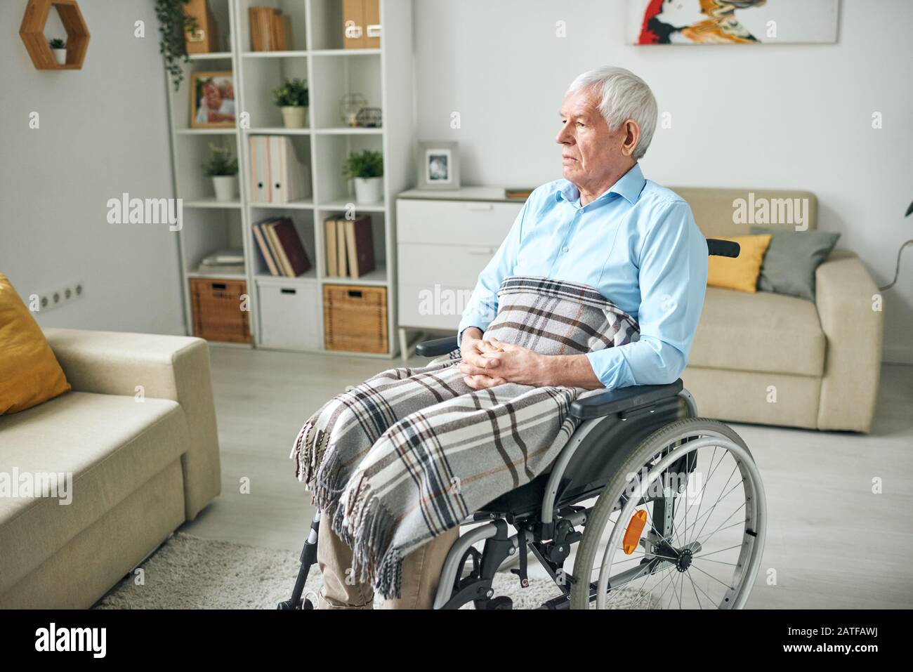 Sad and serene senior disable man sitting in wheelchair by couch at home Stock Photo
