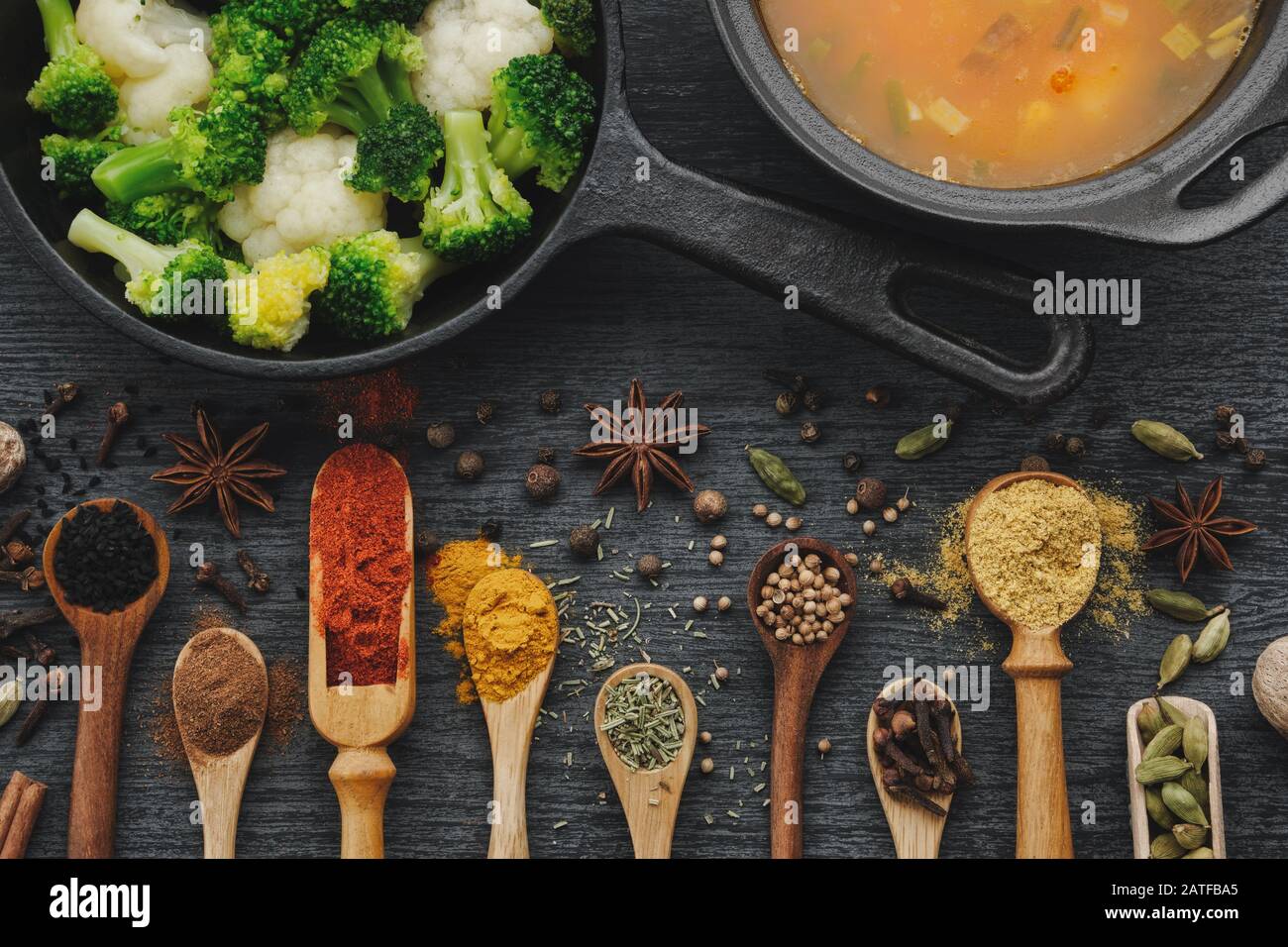 Various aromatic colorful spices and herbs in wooden spoons and scoops. Broccoli and cauliflower on iron frying pan.  Black ceramic saucepan with soup Stock Photo