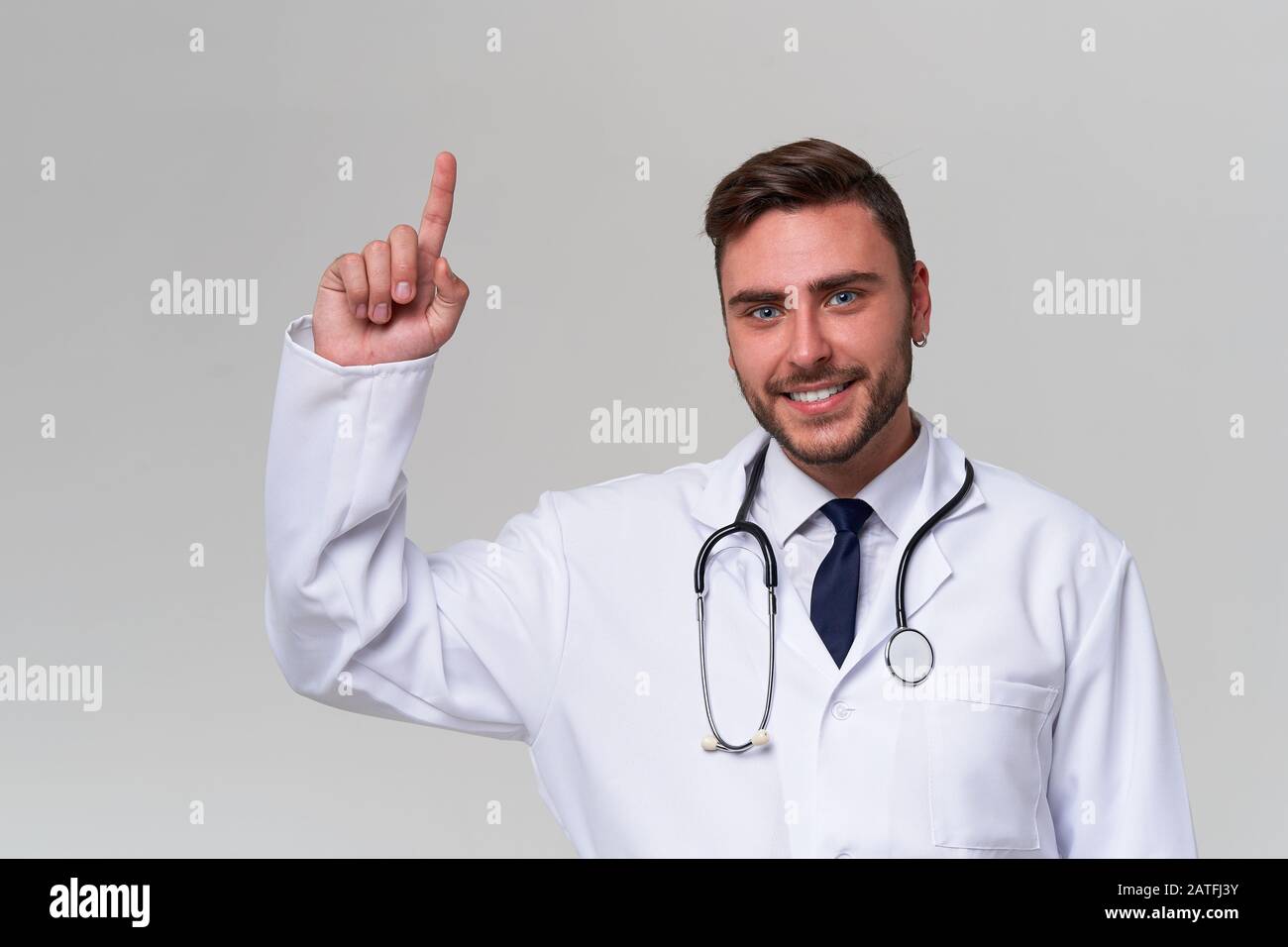 Handsome modern doctor white medical gown stands in studio gray background advertising your product looking at camera and pointing finger away Positiv Stock Photo