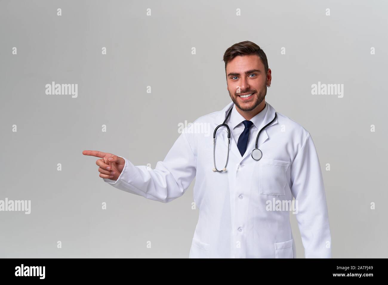 Handsome modern doctor white medical gown stands in studio gray background advertising your product looking at camera and pointing finger away Positiv Stock Photo
