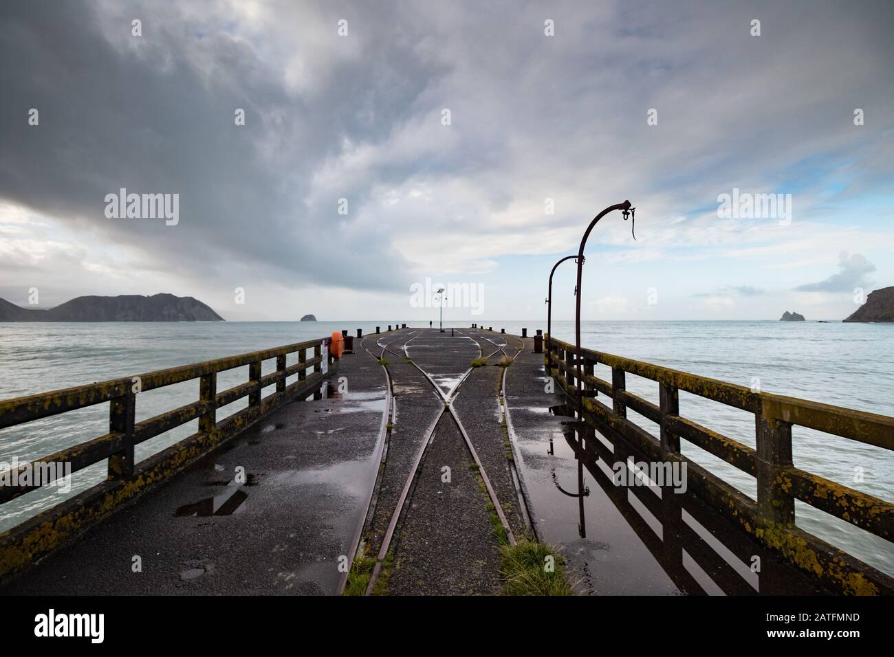 View from the longest wharf (Tolaga Bay) in New Zealand with dramatic clouds and reflections in a puddle along rail tracks and cranes. Stock Photo