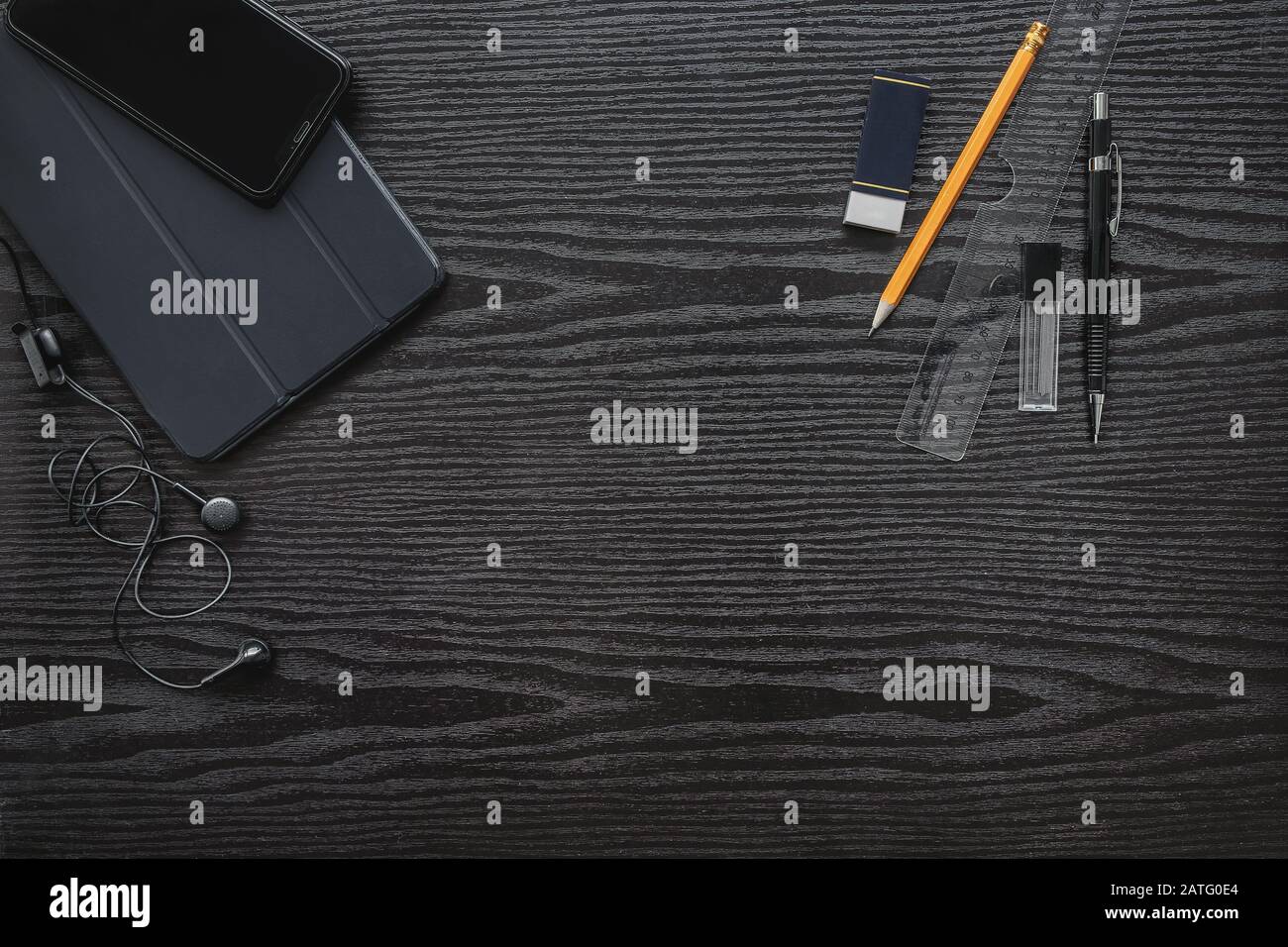 On a black office desk with a wooden texture are a tablet, phone, headphones, and on the other side of the table are stationery Stock Photo