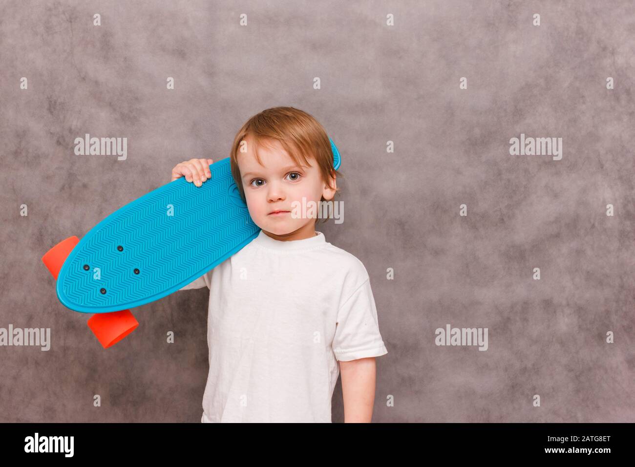 Little boy wearing a casual outfit, hold his skateboard on his shoulder,standing on a gray background. sale concept sports equipment Stock Photo