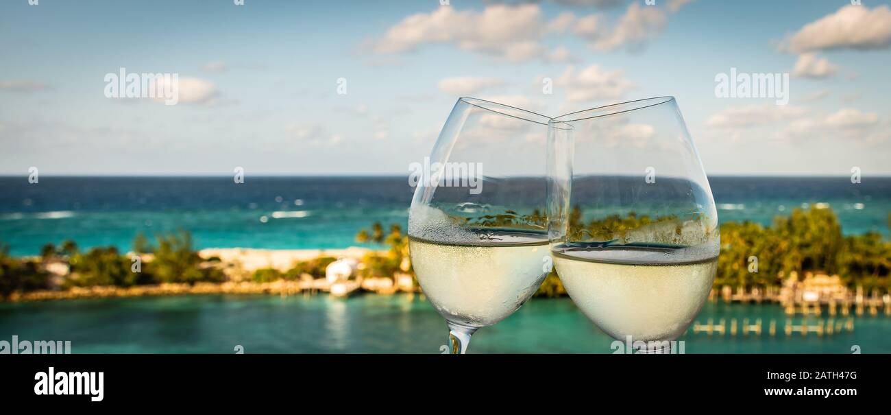 Wide image with a close-up of two clinking glasses of white wine outdoor. Sea and island background at the port of Nassau in the Bahamas. Stock Photo