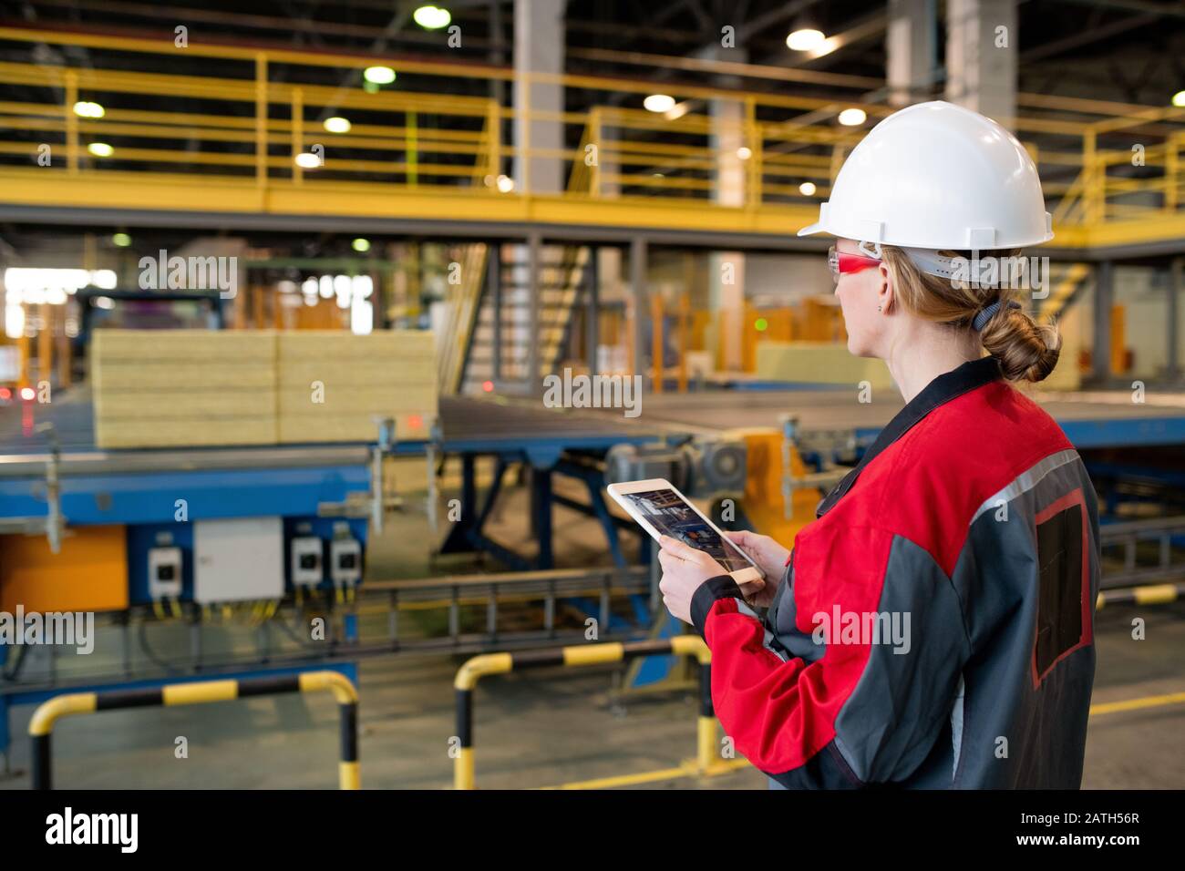 Rear view of busy woman in hardhat and safety goggles using tablet while controlling production line process at factory Stock Photo
