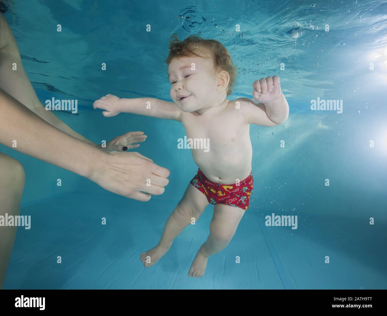Funny red-haired boy plays underwater in a swimming pool, mother holding the child. Stock Photo