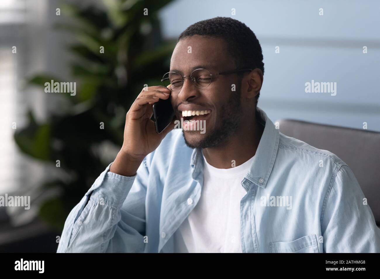 African ethnicity guy laughing during smartphone conversation seated indoors Stock Photo