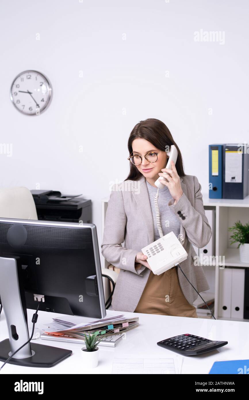 Content attractive young businesswoman in glasses standing at desk and talking on phone in office Stock Photo