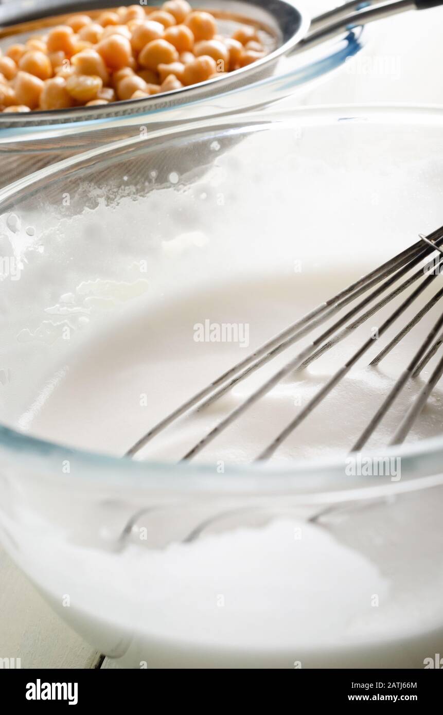Glass bowl of whipped aquafaba (vegan egg white replacer) in the foreground with balloon whisk. Chickpeas (garbanzo beans) straining through metal sie Stock Photo