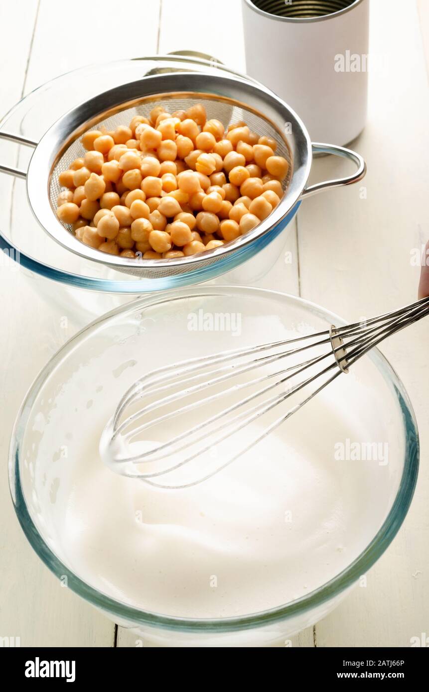 Angled shot of whipped aquafaba in glass bowl with whisk and draining chickpeas in sieve. An empty, unbranded tin sits in background on white painted Stock Photo