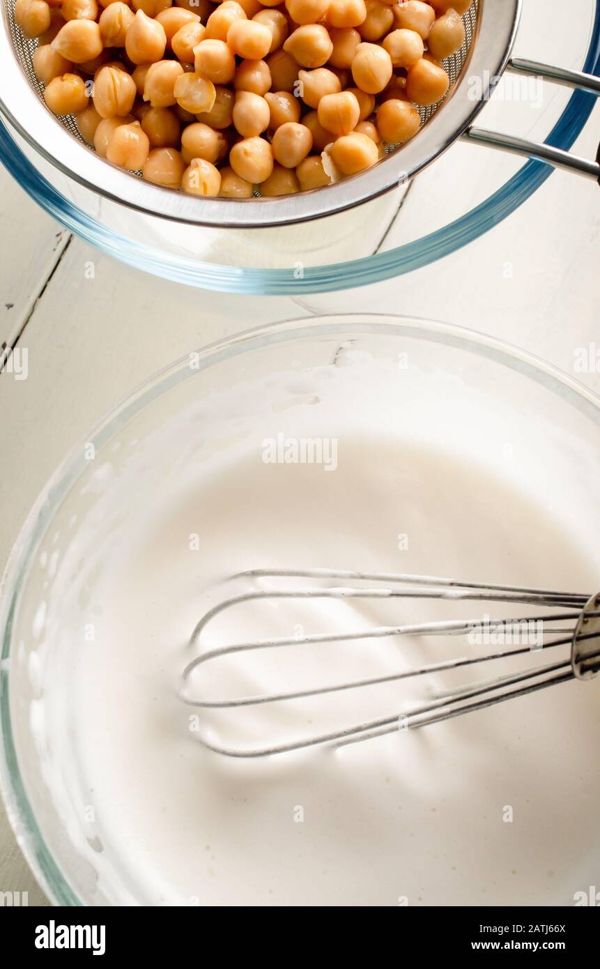 Overhead shot of whipped aquafaba in glass bowl with balloon whisk and chickpeas straining through metal sieve on painted white wood planked table. Stock Photo