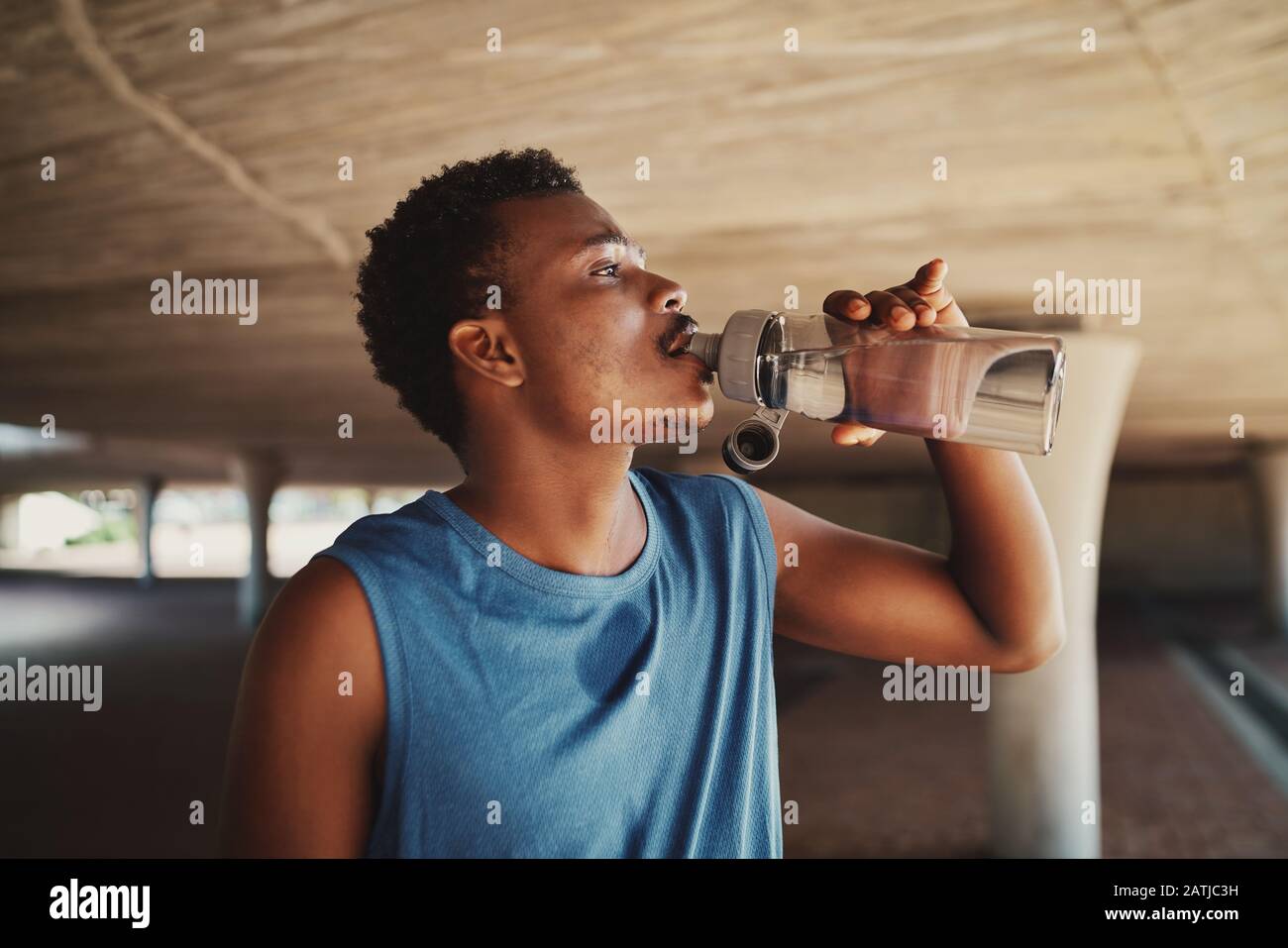 Side view of a young african american man drinking water from bottle standing under the bridge Stock Photo