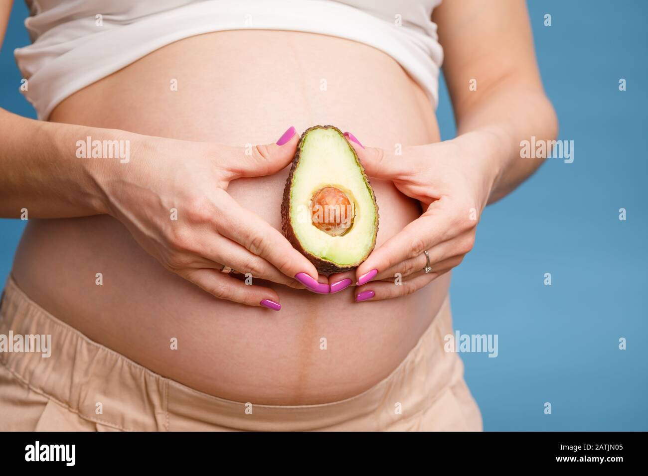 Studio shot of young pregnant woman holding one half of a fresh avocado fruit close to her belly against blue background - healthy nutrition and pregn Stock Photo