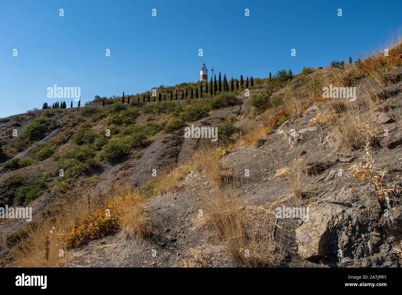 A steep rocky slope with slender cypresses and a lighthouse on a sunny summer day, Crimea. Stock Photo