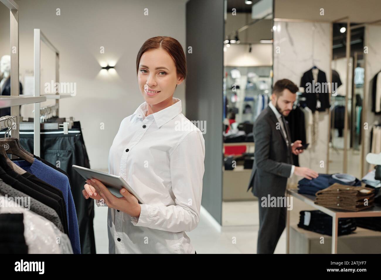 Portrait of smiling beautiful young saleswoman in shirt standing at rack and checking receiving merchandise in clothes shop Stock Photo