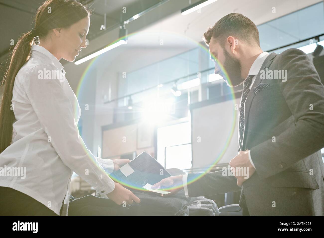 Young woman with ponytail consulting male customer on price issue while showing him jeans in store, lens flare effect Stock Photo