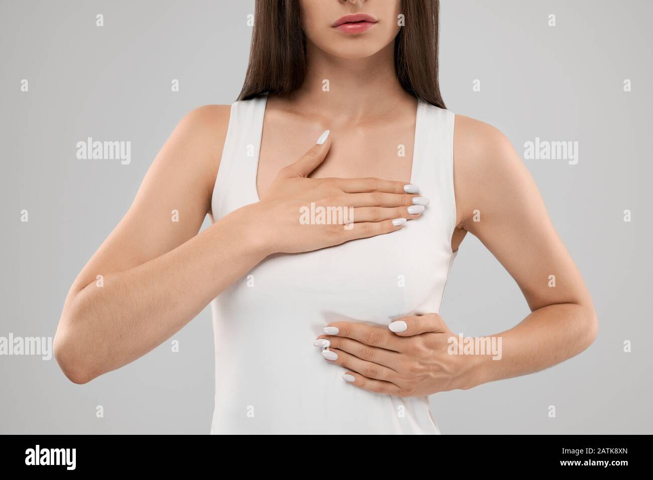 Clolse up of incognito caucasian woman in white shirt posing over gray isolated background and touching chest with two hands because of heartache. Front crop of brunette suffering from pain in heart. Stock Photo