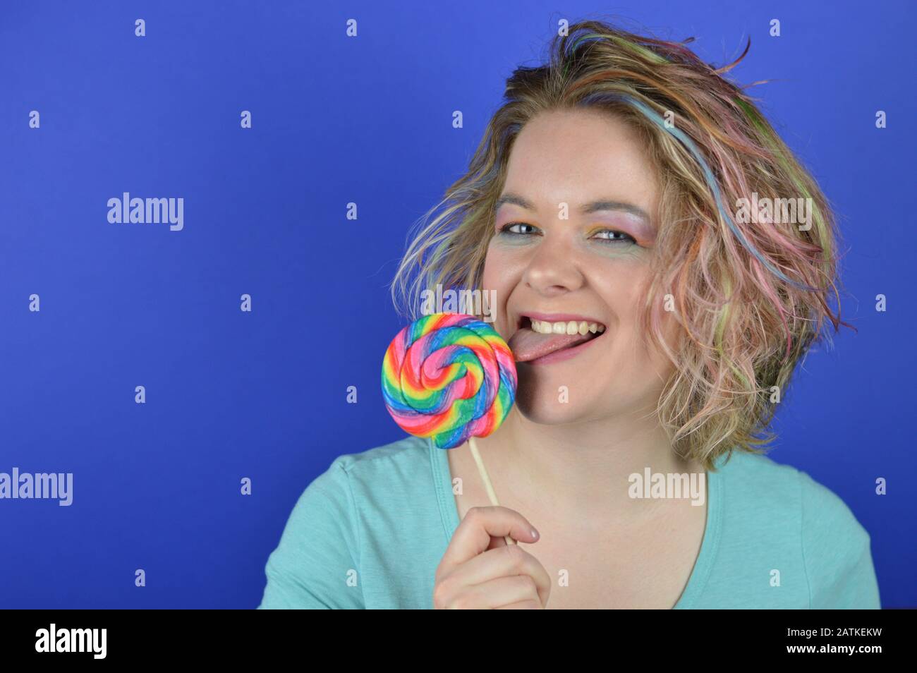 portrait of a young blond woman with colourful hair licking at a big lollipop and looking into the camera Stock Photo