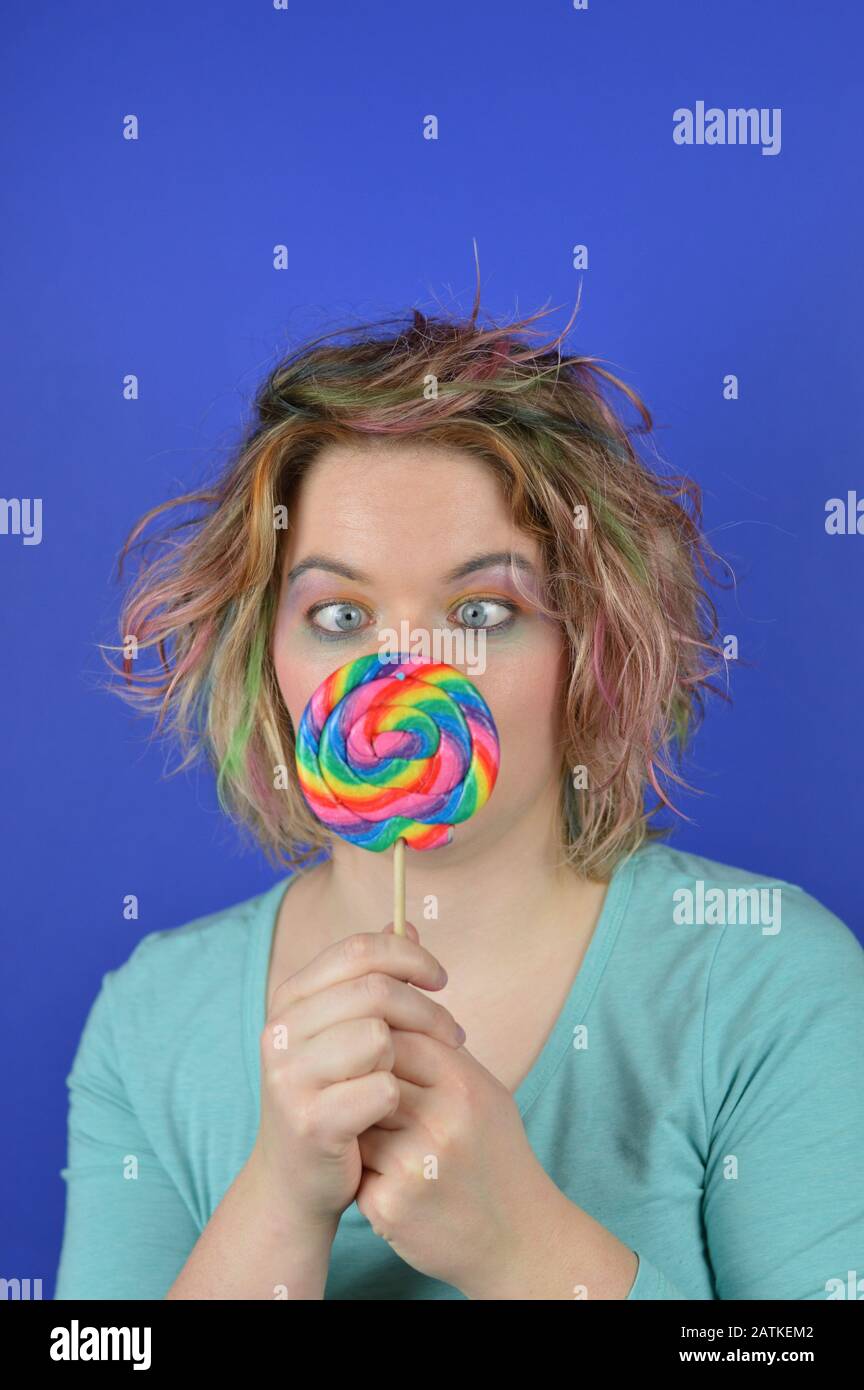portrait of a young blond woman with colourful streaks in the hair and a big sweet multicolored lollipop in her hands squinting and having fun Stock Photo