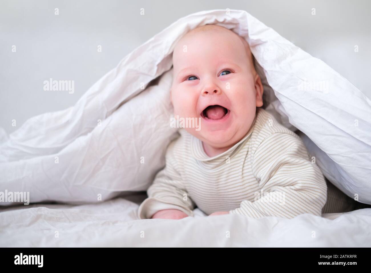 Adorable smiling baby boy under white blanket in bedroom. Newborn child relaxing in bed. Family