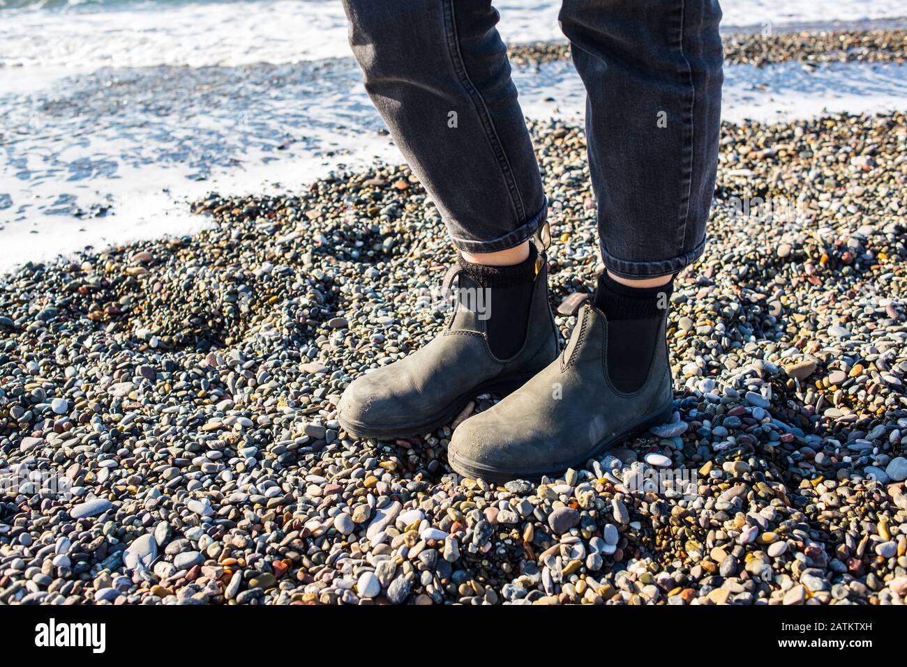 Chelsea boots classic black leather rubber sole. Focus on legs of hipster woman wearing denim black trousers. Shot on gravel beach with sea in backgro Stock Photo
