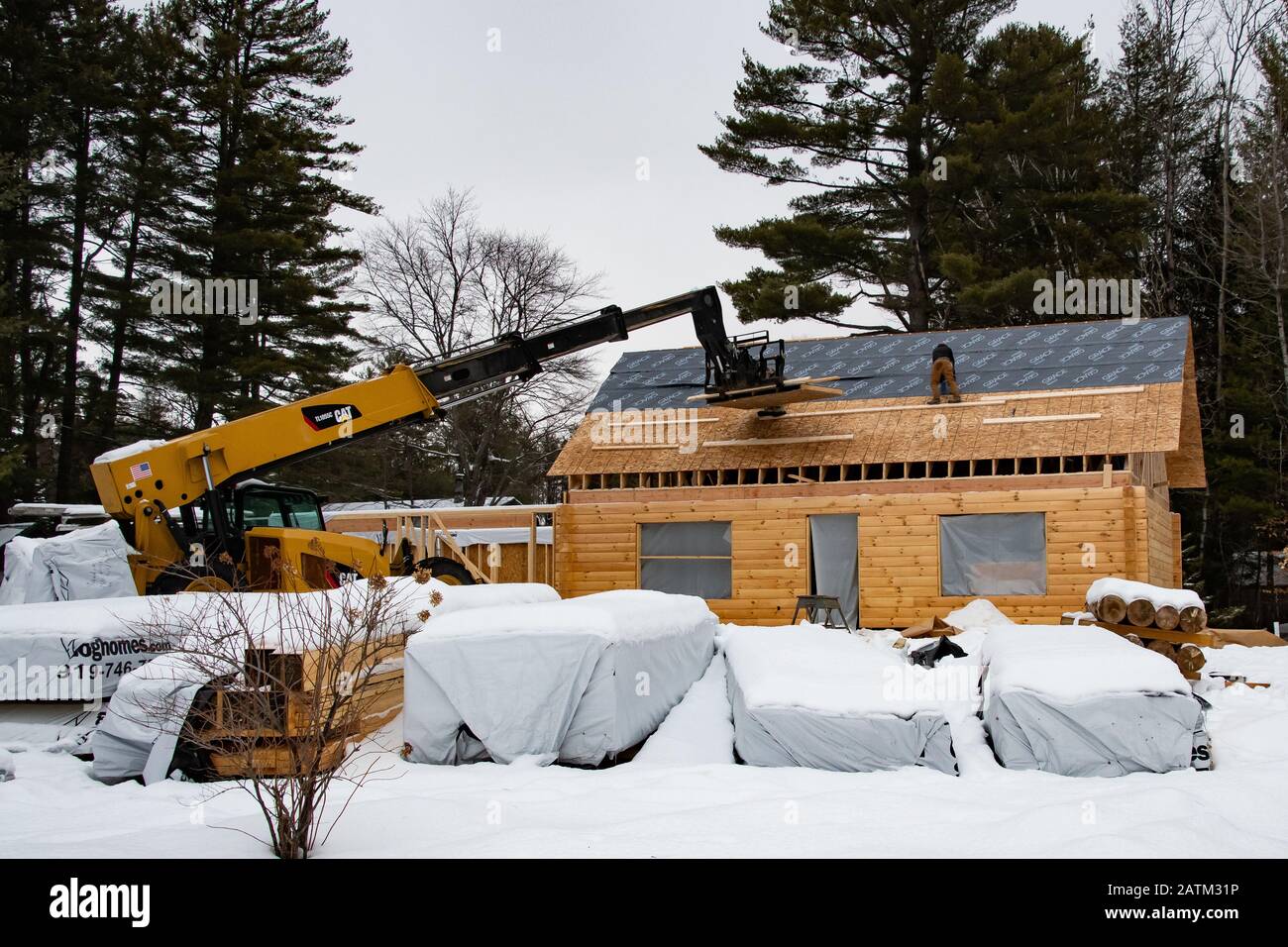 Two construction workers installing a roof covering on a manufactured log home in winter with a Cat TL1055C Telehandler in Speculator, NY USA . Stock Photo
