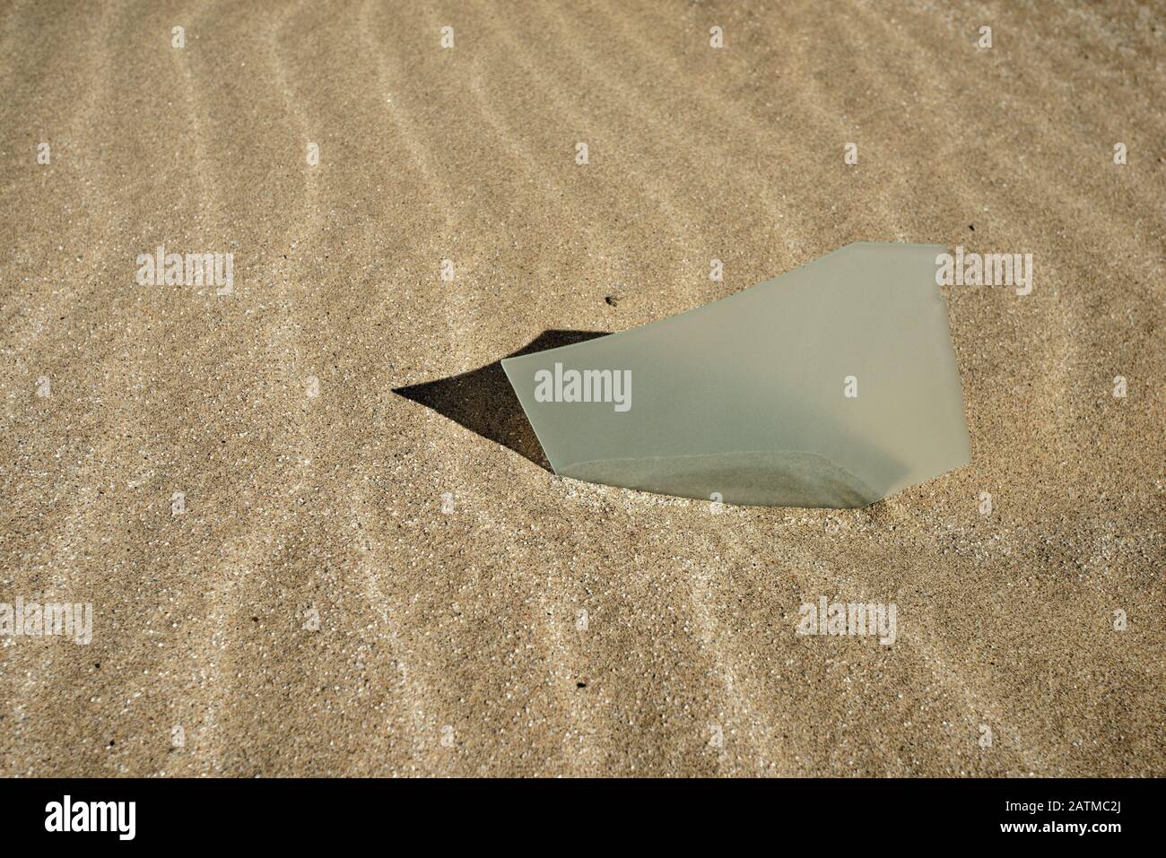 Cutting edge, jagged glass shard embedded in sand with shadow. Stock Photo