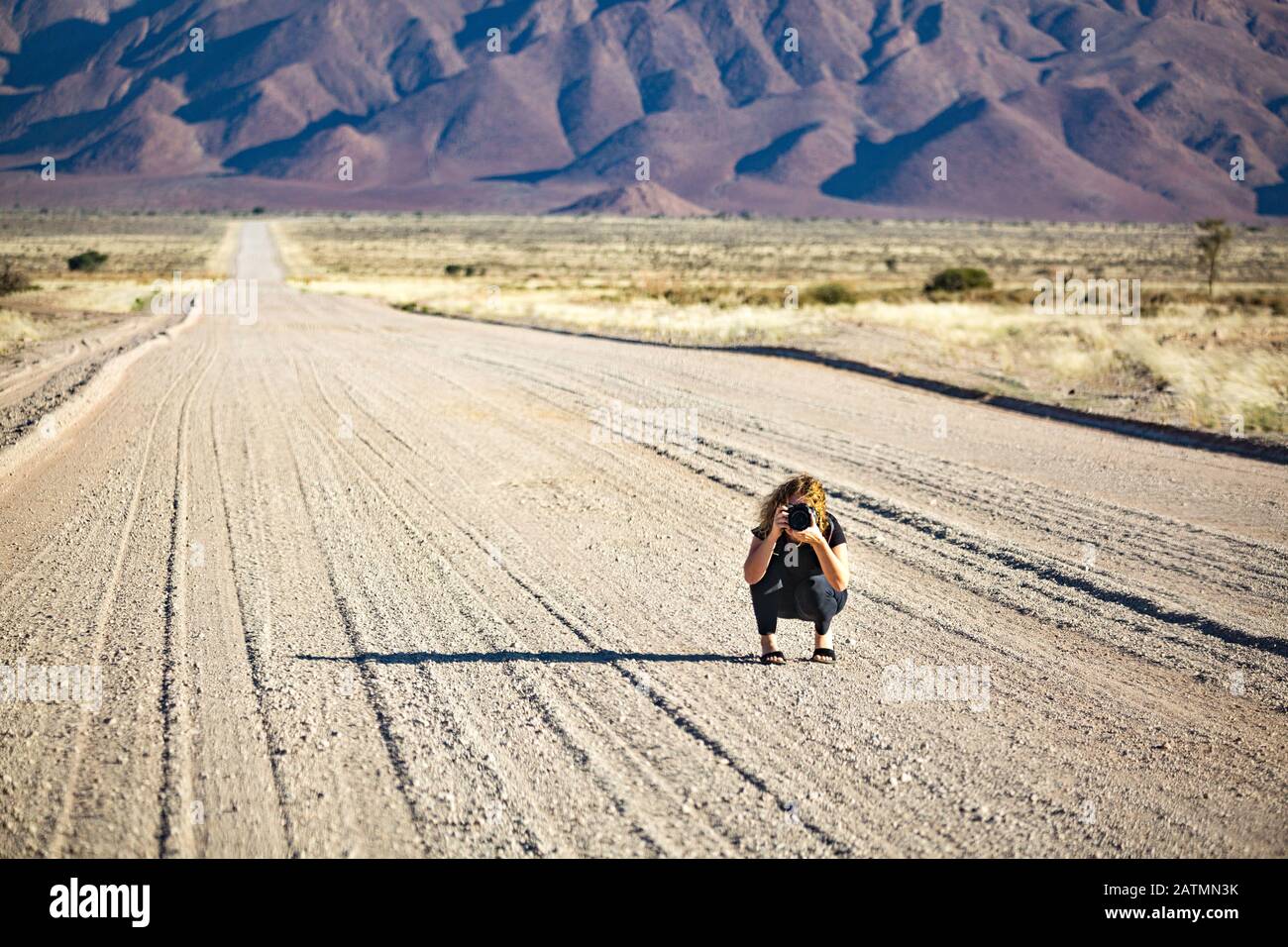 Young woman with a camera squatting in the middle of a gravel road that is leading into a mountainous landscape near Spreetshoogte Pass, Namibia, Afri Stock Photo