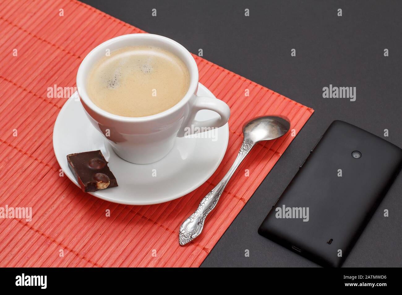 Cup of coffee on saucer with piece of chocolate bar and spoon on bamboo napkin, mobile phone on a black background. Top view. Stock Photo