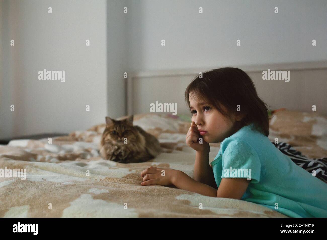 Cute sad little girl watching TV alone in the bedroom with a cat lying on bed, real people, side view Stock Photo