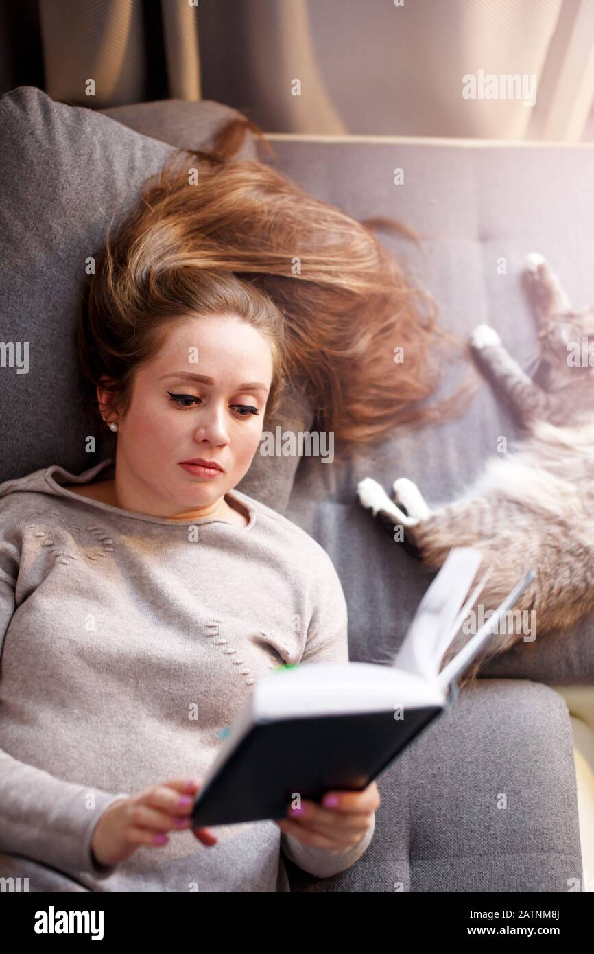 Attractive female with long hair reading interesting book, resting with cat at home, top view Stock Photo