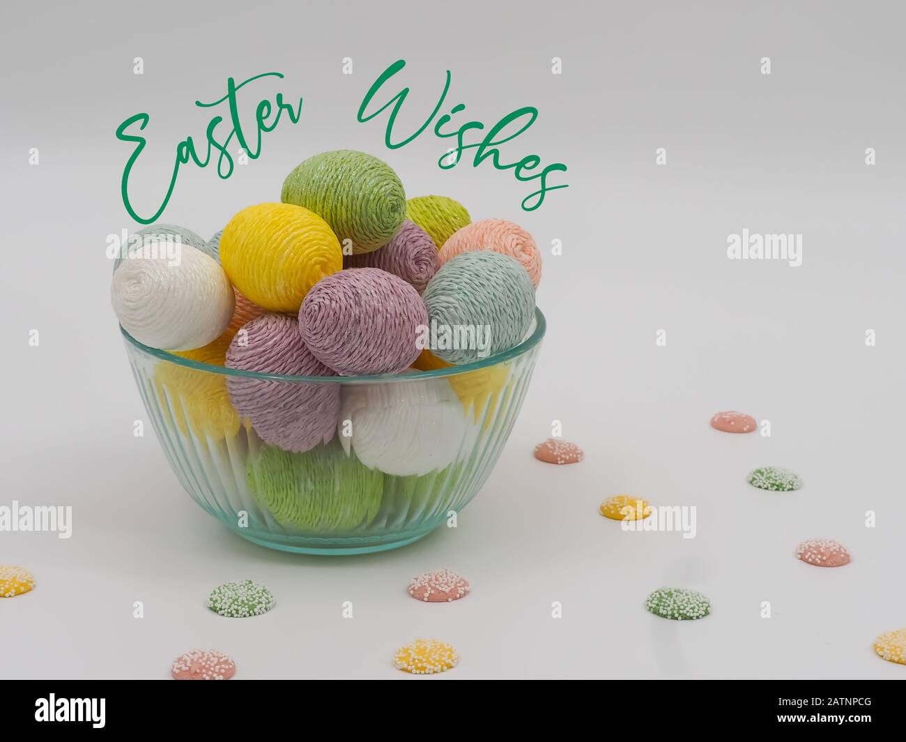 Easter Wishes in green cursive font on a white background with a glass bowl full of string decorated Easter eggs and pastel colored white chocolate ca Stock Photo