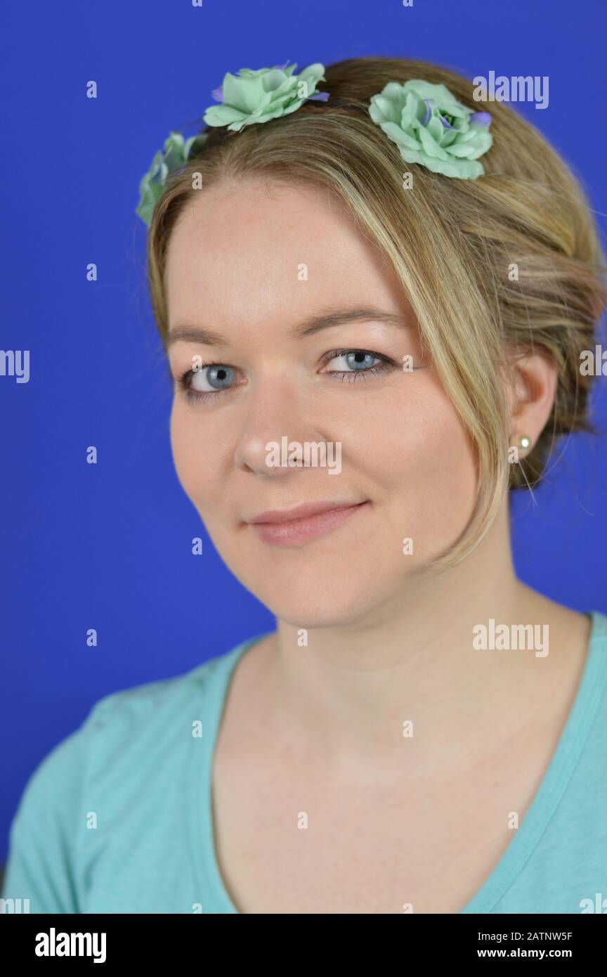 portrait of a pretty young blond caucasian woman with updo hair and cyan flowers on a hair circlet smiling in front of blue background Stock Photo