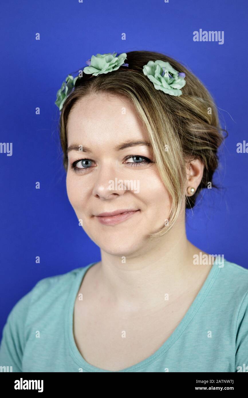 portrait of a pretty young blond caucasian woman with updo hair and cyan flowers on a hair circlet smiling in front of blue background Stock Photo