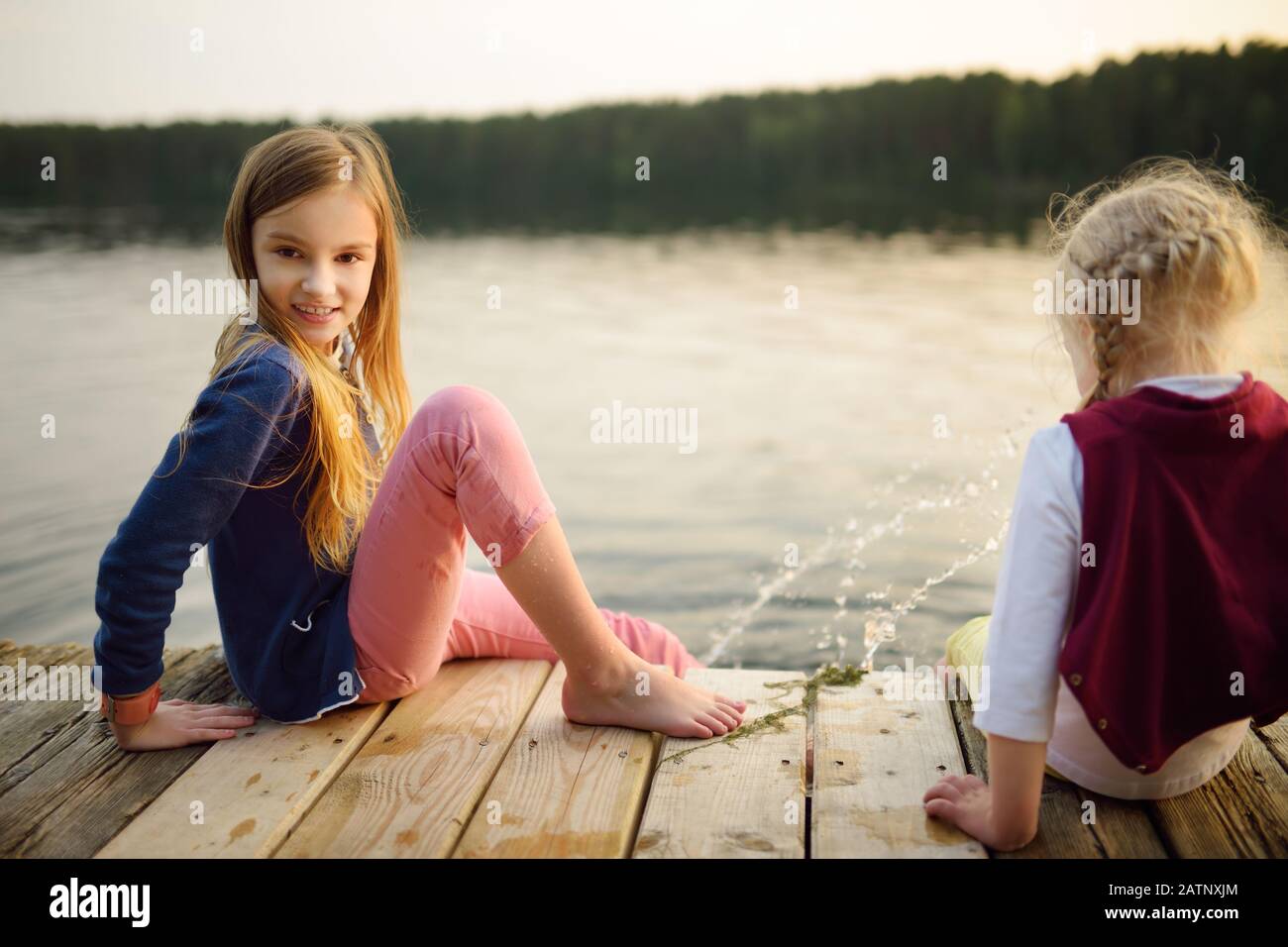 Two cute little girls sitting on a wooden platform by the river or lake dipping their feet in the water on warm summer day. Family activities in summe Stock Photo