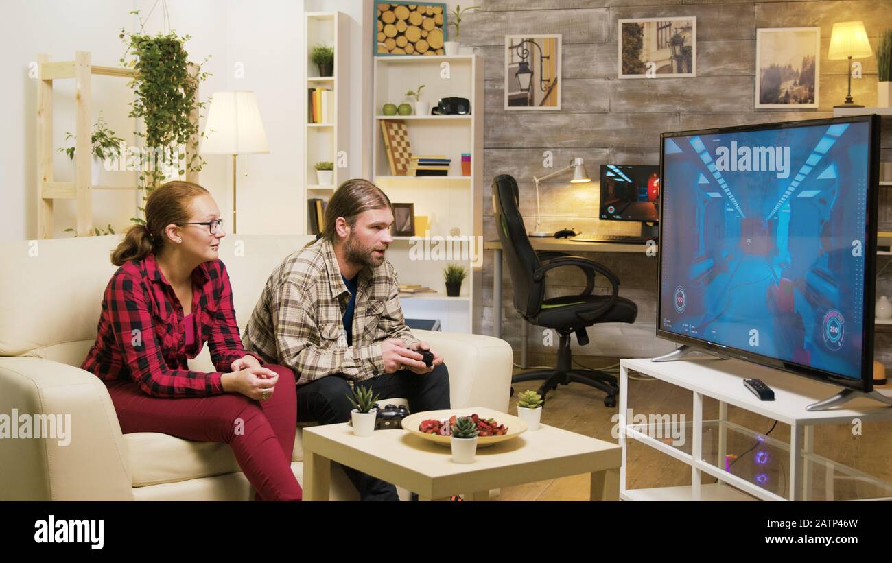 Cheerful girlfriend talking with her boyfriend while he's playing video games on tv using wireless joystick. Stock Photo