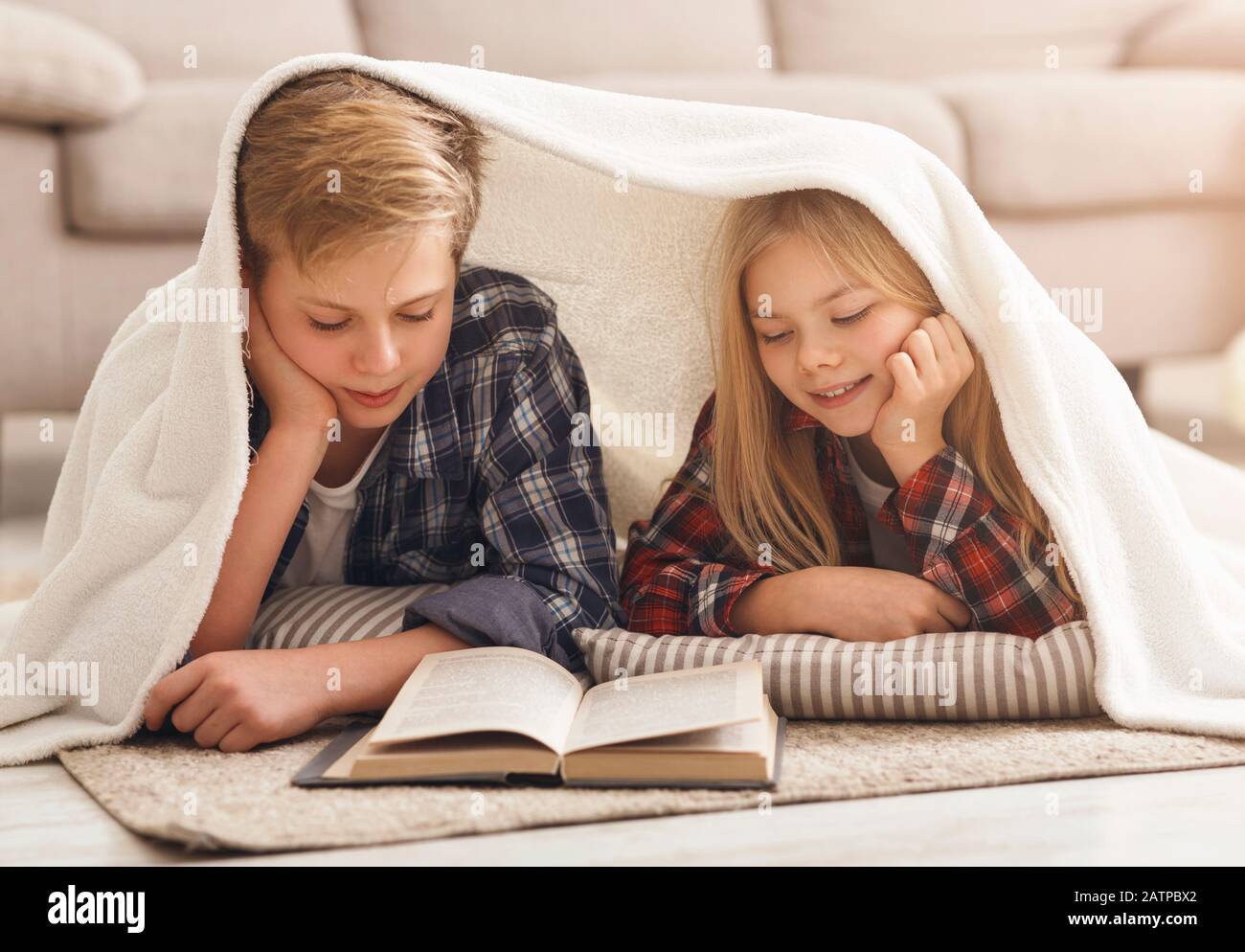 Brother And Sister Reading Book Covered With Blanket At Home Stock Photo