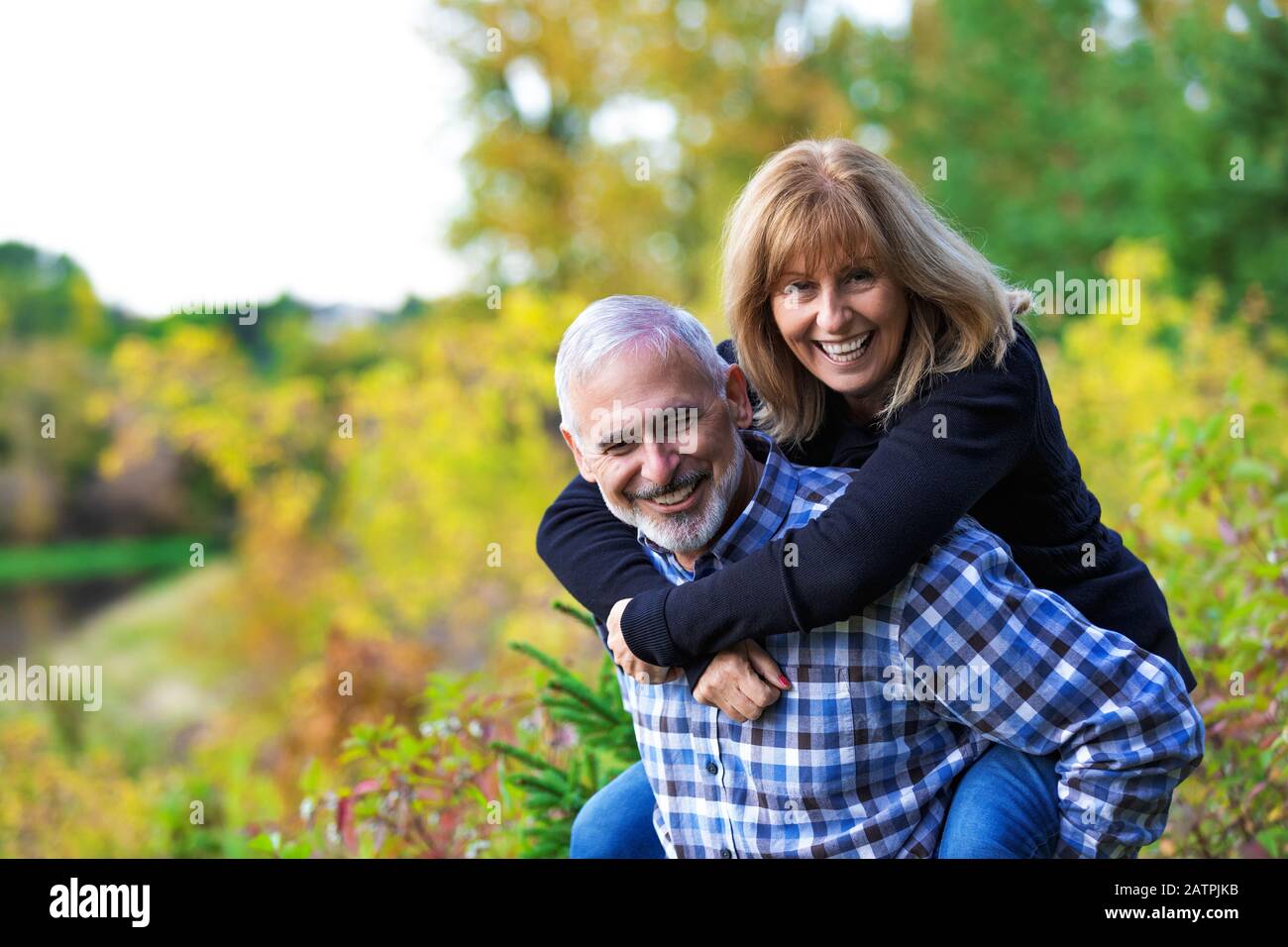 A mature huband having fun and giving his wife a piggy back ride on a path along a river in a city park on a warm fall evening Stock Photo