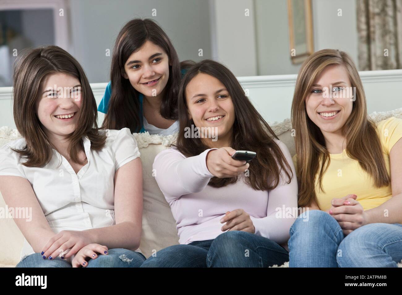 Four teenage girls sitting on a couch at home watching TV and changing the channel with the remote control Stock Photo