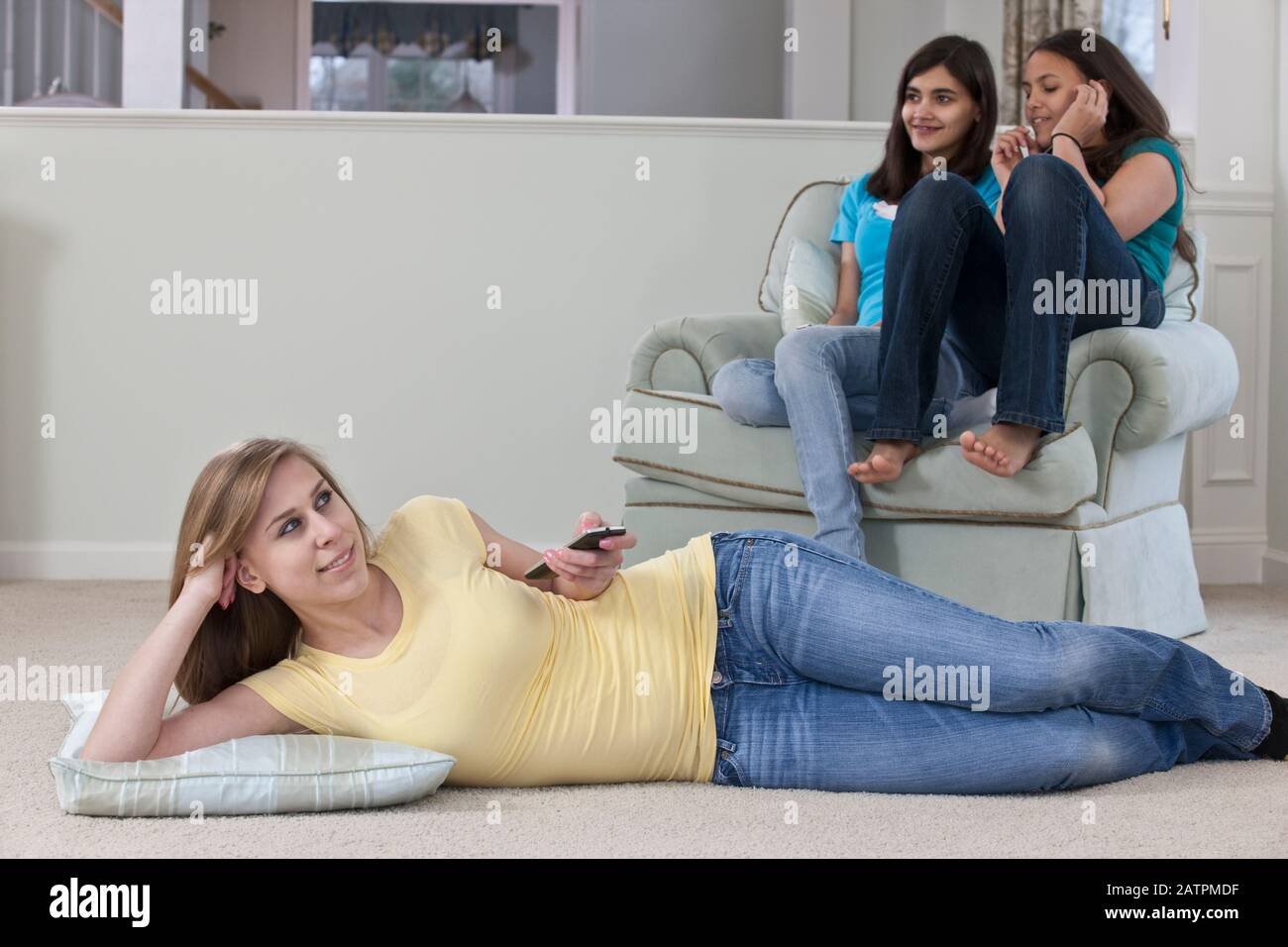 Three teenage girls at home together, one is watching TV and changing the channel with the remote control and the others are in a chair listening t... Stock Photo