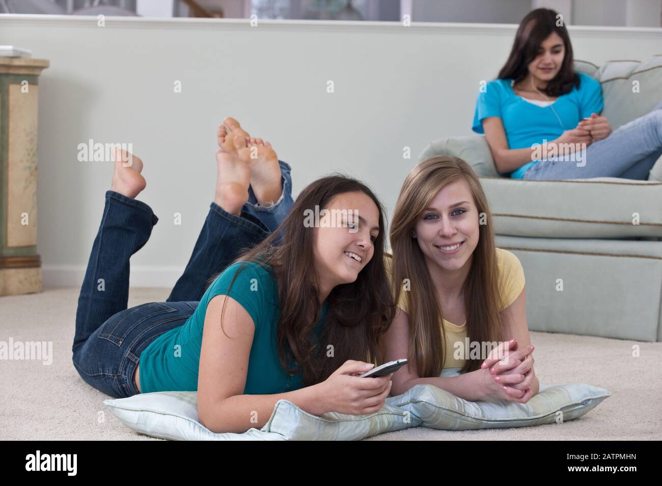 Three teenage girls at home together, two are watching TV and changing the channel with the remote control and the other is in a chair listening to... Stock Photo