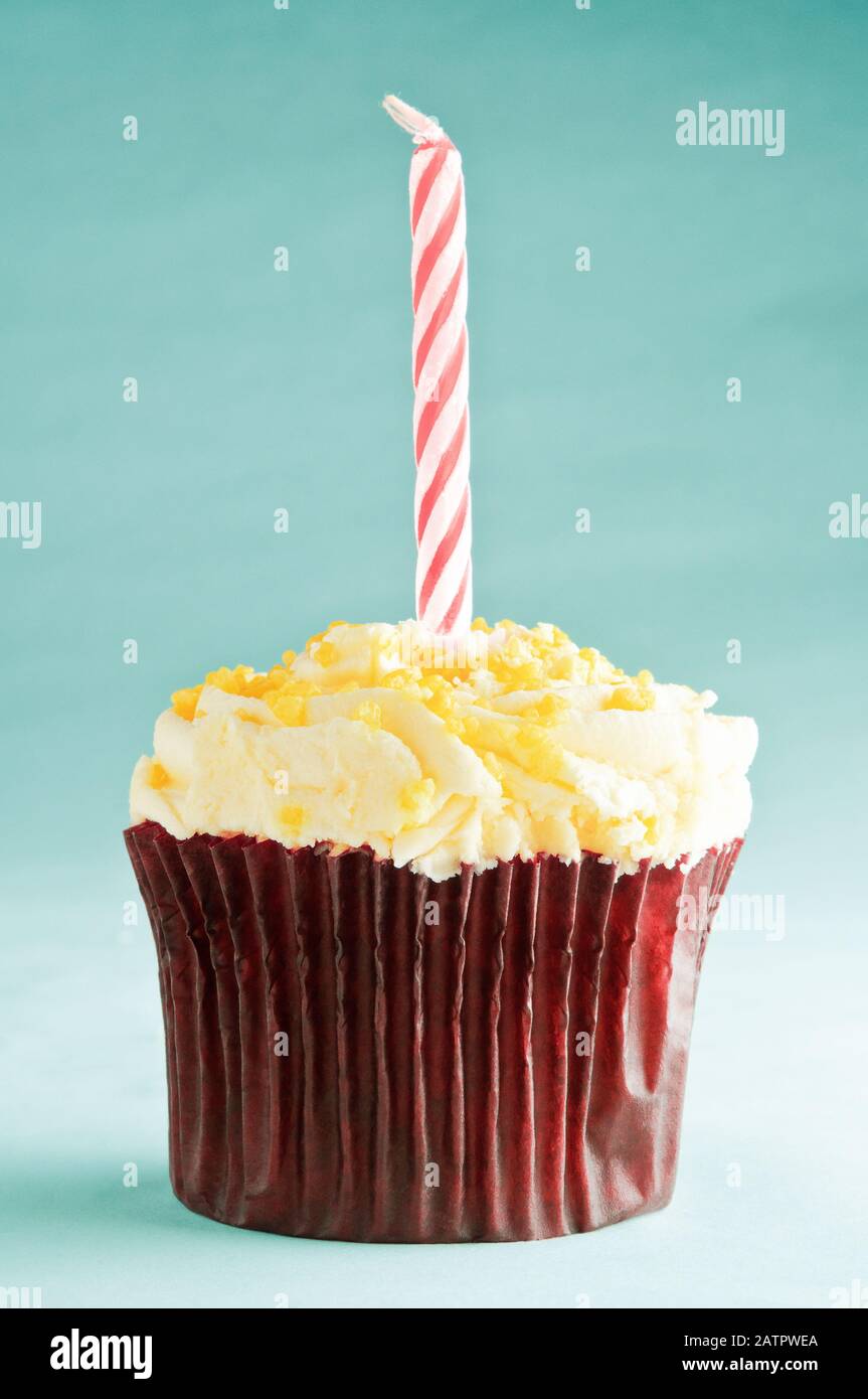 A single birthday cupcake with one striped red and white candle in brown paper case with yellow buttercream icing and sprinkles. Turquoise background. Stock Photo