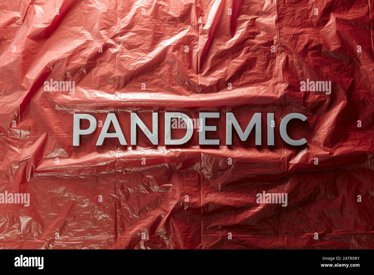 the word pandemic laid with silver letters on red crumpled plastic film background in flat lay composition at center with dramatic cold light Stock Photo